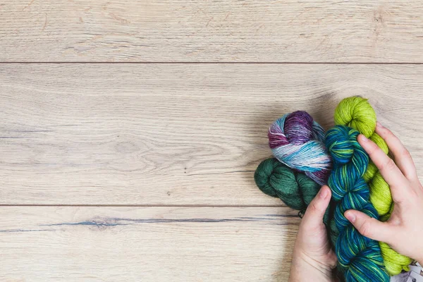 Flat lay of human hands holding beautiful skeins in purple and green tones over a wooden table and copy space