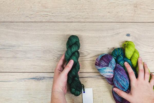 Flat lay of human hands choosing beautiful skeins in purple and green tones with blank label for mock up over a wooden table