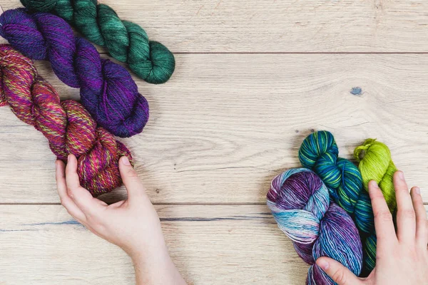 Flat lay of human hands choosing beautiful skeins in purple and green tones over a wooden table