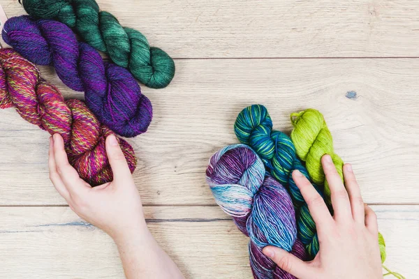 Flat lay of human hands choosing beautiful skeins in purple and green tones over a wooden table
