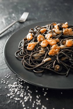 Deniz tuzu ve gümüş çatalın yanında sepya mürekkebi ve karidesli siyah makarna tabakası ve siyah desenli arka plan.