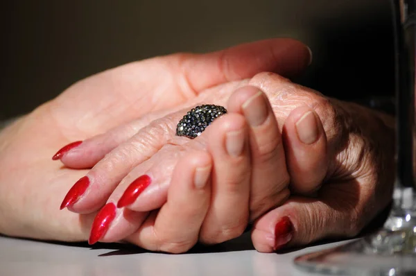 Adult daughter holding mother wrinkled old hand wearing a ring and red nail polish. Scene partially lit with a dramatic soft effect. Hands on the table/ adult daughter holding her mother hand/