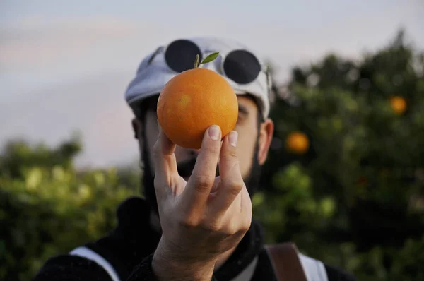 Vintage man holding an orange at the height of the face/ covering the face with an orange/ Man wearing rounded sunglasses and gray beret/ showing an orange with the right hand