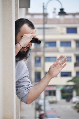 Genç bir adam, dairesinin camından dışarı sarkmış dürbünle ailesine el sallıyor. İspanyol kasabasında karantina zamanında dürbünle pencereden izleyen bir adam var.