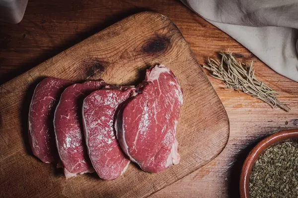 Cuts of beef on wooden board along with oregano, rosemary and a knife