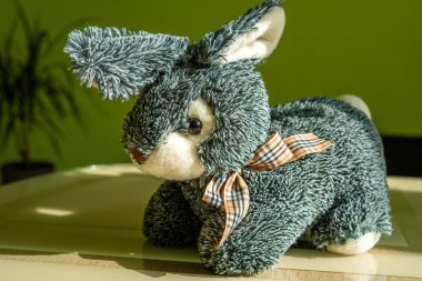 İzole edilmiş gri tüylü oyuncak tavşan portresi. Tekstil kareli boyun bandı açık kahverengi yüzeyde duruyor. Sıcak yeşil arka planda.