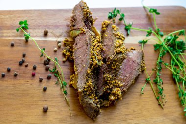 Mutfaktaki ahşap bir kesim tahtasının üzerinde taze yeşil kekik ve tam biberle çevrili tam tahıllı hardalla kaplı pembe merkezli fırında sığır eti dilimleri yakından görülüyor.