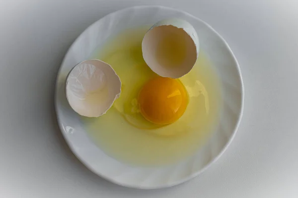Top down view of one cracked egg with yellow yolk surrounded by egg shells is laying on a white plate on white background