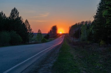 Parlak turuncu günbatımını baharda ormanda yolun üzerinde gösteren kırsal bir manzara.