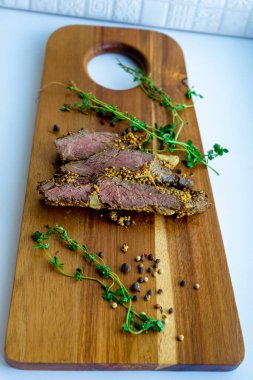 Mutfaktaki ahşap bir kesim tahtasının üzerinde taze yeşil kekik ve tam biberle çevrili tam tahıllı hardalla kaplı pembe merkezli fırında sığır eti dilimleri yakından görülüyor.