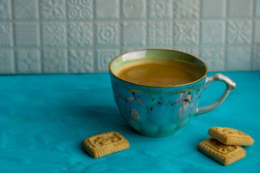 Mutfaktaki beyaz seramik fayanslara karşı turkuaz yüzeyde duran siyah kahve ve küçük kurabiyelerle dolu fincanın yakın görüntüsü.