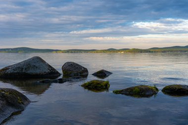 Büyük gri taşlar baharda bulutlu bir havada bir gölün suyunda yatıyor.