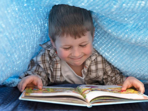 A boy under a blanket reads a book, a boy with a book at home