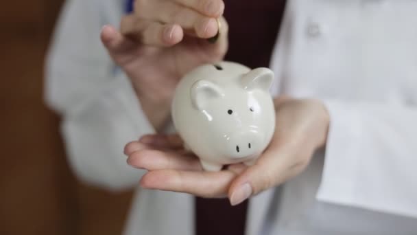 Primer plano de las mujeres doctor mano poner moneda en alcancía. Concepto de seguro médico — Vídeo de stock