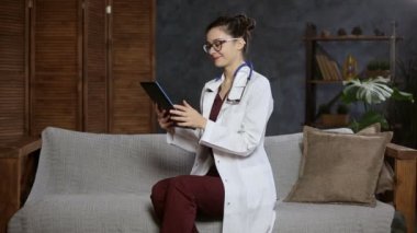 Kadın doktor kanepede oturuyor ve tabletle video konferansı yapıyor. Online danışman hasta
