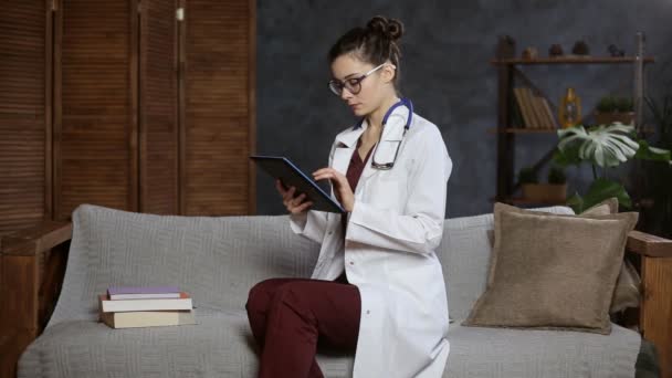 Портрет жінки-лікаря зі стетоскопом за допомогою планшетного комп'ютера на дивані — стокове відео