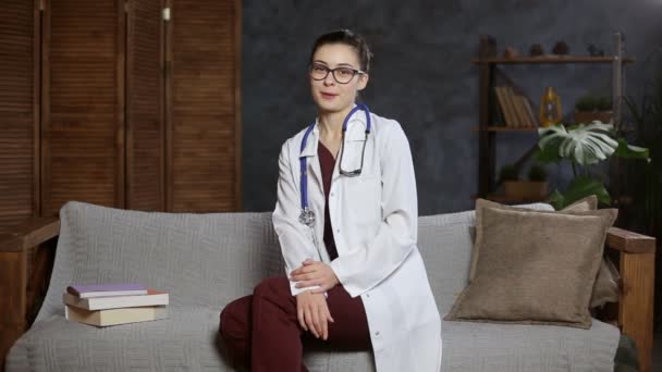 Professionele vrouw arts met stethoscoop en bril praten met camera zitten op de bank. — Stockvideo
