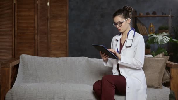 Retrato de una doctora con estetoscopio usando tableta en el sofá, espacio para copiar — Vídeo de stock