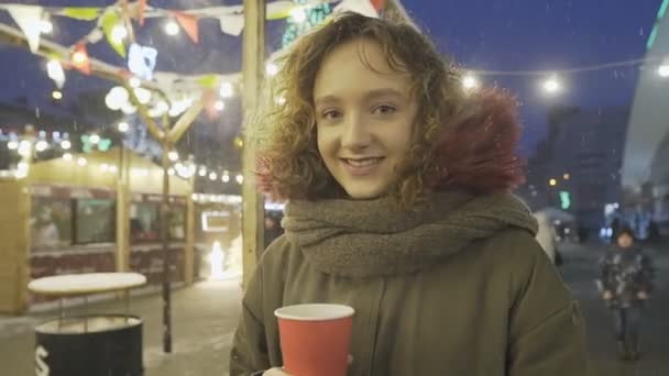 Portret młodej dziewczyny z kręconymi włosami pozowanie z filiżanką kawy na jarmarku bożonarodzeniowym. — Wideo stockowe