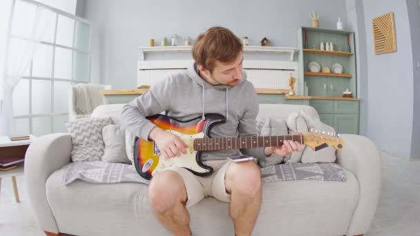 Férfi a kanapén játszik az elektromos gitáron okostelefonnal otthon. Online tanulási koncepció. — Stock videók