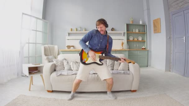 Wesoły młody człowiek ze słuchawkami grający na gitarze elektrycznej w salonie w domu — Wideo stockowe