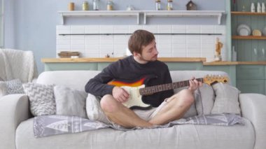 Evdeki kanepede elektro gitar çalan genç adam.