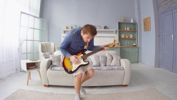 Joven hombre divertido con auriculares saltando y tocando la guitarra eléctrica en casa — Vídeo de stock