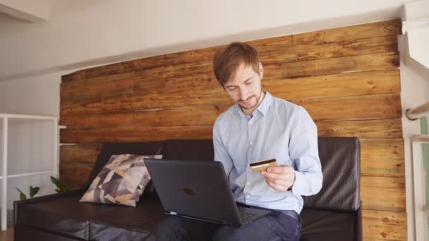 Молодой бизнесмен с ноутбуком и кредитной картой делает покупки онлайн в мансардной квартире . — стоковое видео