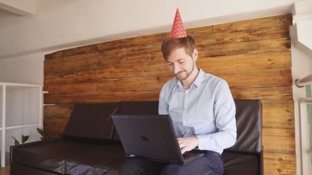 Hombre sonriente guapo con gorra de cumpleaños usando el ordenador portátil, buscando regalos — Vídeos de Stock