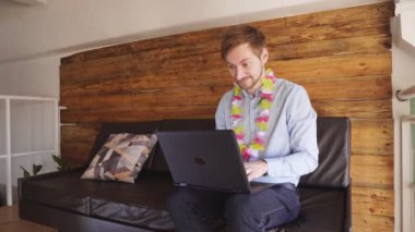 Koltukta dizüstü bilgisayarla oturan ve başarıyı kutlayan Hawaii kolyeli mutlu genç adam.