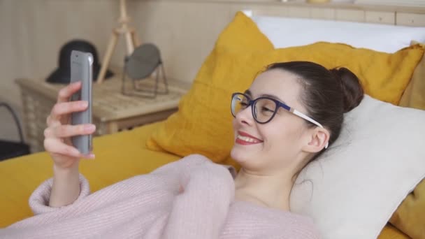 Женщина машет во время видеозвонка на смартфон, лежащий на кровати дома — стоковое видео
