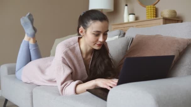 ソファに横になりながらノートパソコンで働く若い女性の肖像画。オンライン教育 — ストック動画