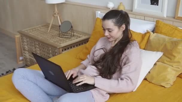 Молодая привлекательная улыбчивая женщина, лежащая на кровати, работает над новым проектом с ноутбуком дома — стоковое видео