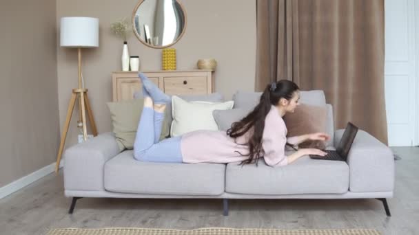 Молодая брюнетка женщина расслабляется с ноутбуком, лежа на диване в гостиной, работая из дома — стоковое видео
