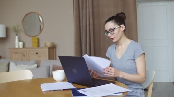 Mujer joven con gafas leyendo documentos mientras trabaja en un portátil en casa. Trabajo a distancia o educación en línea. Quédate en casa mientras estás en cuarentena contra el coronavirus . — Vídeos de Stock