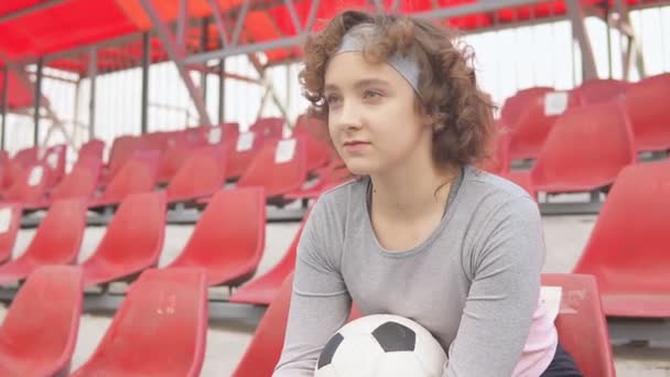 Молодая девушка-подросток с футбольным мячом сидит на сиденьях на футбольном стадионе и мечтает — стоковое видео