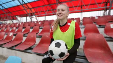 Futbol topuyla gülümseyen genç futbolcu kız futbol stadyumunda oturuyor ve hayal kuruyor.