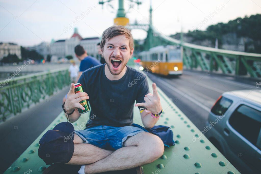 Young cheerful bearded man sitting on bridge in Budapest and holding can of beer, happily looking in camera