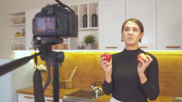 Ευτυχισμένο νεαρό κορίτσι κρατώντας μήλα και καταγράφοντας το επεισόδιο του blog της σχετικά με την υγιεινή διατροφή, ενώ στέκεται στην κουζίνα στο σπίτι — Αρχείο Βίντεο