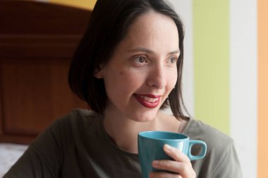 Genç, doğal, güzel bir kadın, huzurlu ve mutlu bir ifadeyle rahat bir sabahta evde kahve içiyor..