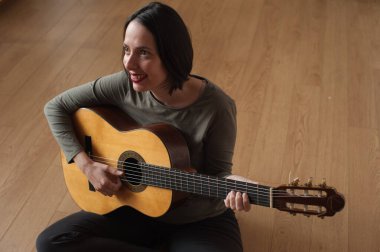 Evde gitar çalan genç ve güzel bir kadın. Ahşap bir arka planda doğal ve rahatlatıcı bir ifadeyle..