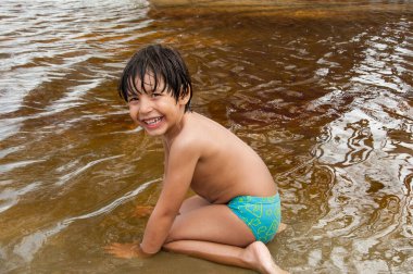 Nehirde mayo giymiş, dizlerinin altında oturan küçük Brezilyalı çocuk kameraya bakıyor ve gülümsüyor..