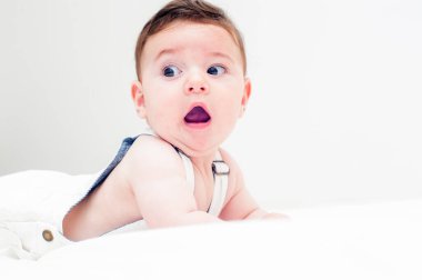 Beyaz tulumlu güzel bir bebek beyaz bir çarşaf ve beyaz bir arka plan içinde karnının üzerinde yatıyor. Ağzı açık ve komik bir ifadesi var..