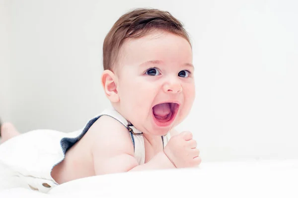 cute baby smiling in white overalls lying on his stomach in a white sheet and a white background.