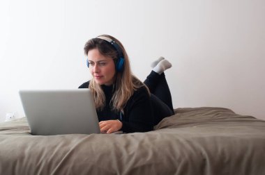 Sarışın kadın bilgisayarı kullanarak yatağa uzanıyor. Kulaklık kullanıyor.
