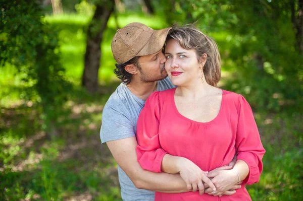 Parkta kucaklaşan bir çiftin ön görüntüsü. Kız kameraya bakıyor ve adam onu yanağından öpüyor..