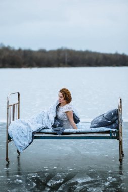 Gecelik ve battaniyeli bir kız, donmuş mavi gölde bir yatakta uyanıyor.
