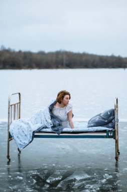 Gecelik ve battaniyeli bir kız, donmuş mavi gölde bir yatakta uyanıyor.