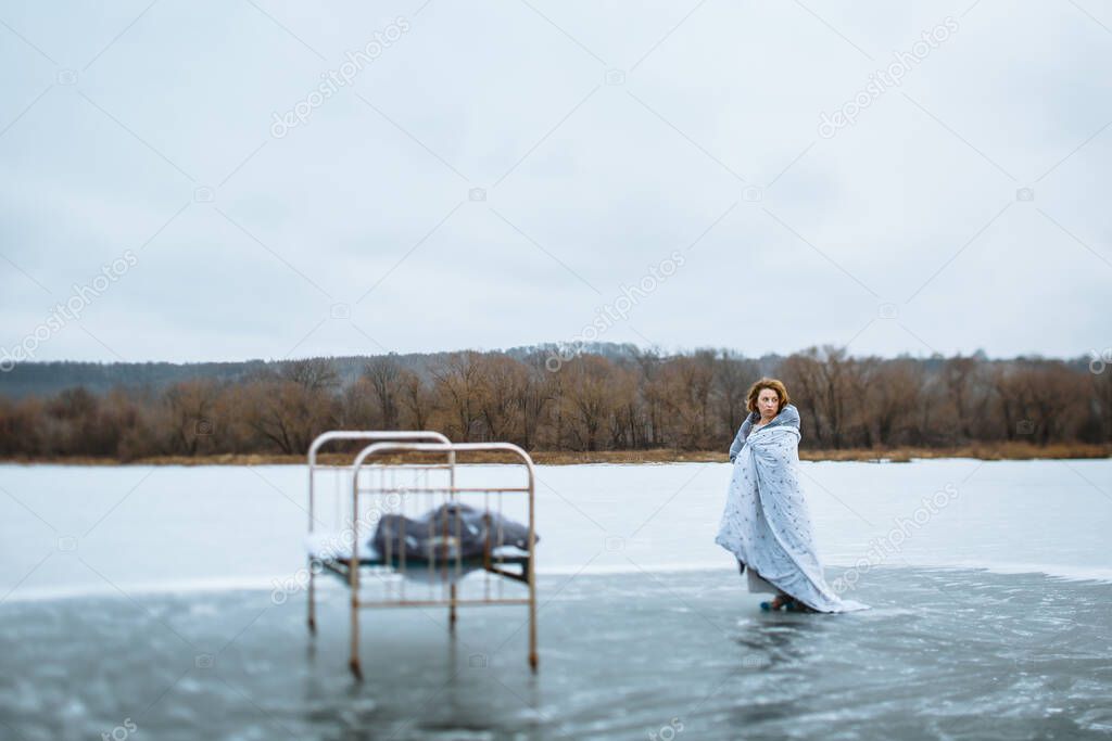 A girl in a nightgown and a blanket standing on the ice of a blue lake, next to an iron bed