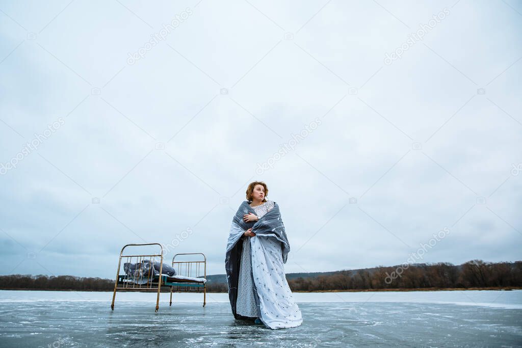 A girl in a nightgown and a blanket standing on the ice of a blue lake, next to an iron bed